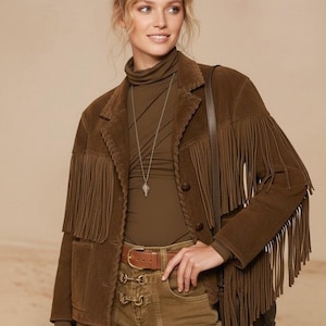 May include: A brown suede jacket with fringing on the sleeves and front. The jacket is worn with a brown turtleneck, matching trousers, and a brown belt. A silver necklace and a brown shoulder bag complete the outfit.