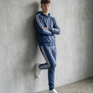 Op de afbeelding: Een marineblauw trainingspak met witte strepen op de mouwen en broekspijpen. Het pak bestaat uit een hoodie met rits en een bijpassende broek. De persoon draagt witte sneakers. De outfit is gemaakt van een glanzend materiaal.