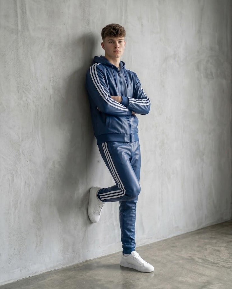 Op de afbeelding: Een marineblauw trainingspak met witte strepen op de mouwen en broekspijpen. Het pak bestaat uit een hoodie met rits en een bijpassende broek. De persoon draagt witte sneakers. De outfit is gemaakt van een glanzend materiaal.