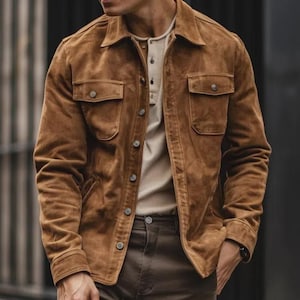 Puede incluir: Chaqueta de ante marrón con dos bolsillos en el pecho y cierre de botones. La chaqueta se lleva sobre una camiseta Henley de color claro y se combina con pantalones marrones. La chaqueta tiene un estilo clásico e informal.