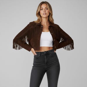 May include: A brown suede jacket with fringe detailing on the sleeves and chest. The jacket has a button-up front and a collar. The model is wearing a white crop top and black jeans. The jacket is a cropped style.