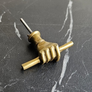 Peut inclure: Une poignée de meuble en laiton en forme de main agrippant une barre. La quincaillerie a une tige filetée pour le montage. La finition est de couleur or antique chaud. La poignée est posée sur une surface sombre et marbrée.