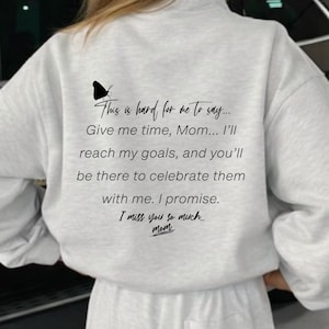 Puede incluir: Conjunto de sudadera y pantalones cortos de color gris claro. La parte trasera de la sudadera presenta una mariposa negra y un mensaje sincero: "Es difícil para mí decir... Dame tiempo, mamá... Alcanzaré mis metas... Te echo mucho de menos, mamá."