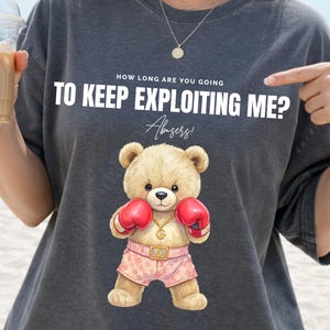 Puede incluir: Camiseta gris oscuro con el texto "HOW LONG ARE YOU GOING TO KEEP EXPLOITING ME?" y un oso de peluche de dibujos animados con guantes de boxeo y pantalones cortos. El oso tiene un collar de oro. La camiseta la lleva una persona.