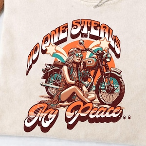Puede incluir: Camiseta color crema con un gráfico retro. El diseño presenta a una mujer con gafas de sol junto a una motocicleta, con el texto "NO ONE STEALS MY PEACE" en naranja y blanco, con estrellas y un fondo circular.