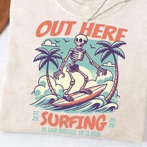 Puede incluir: Camiseta color crema con un gráfico de un esqueleto surfeando. El diseño incluye palmeras, olas y el texto "OUT HERE SURFING in the middle of a war". La camiseta tiene una etiqueta Comfort Colors.