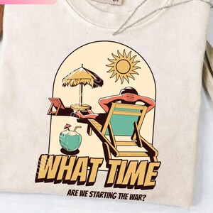 Puede incluir: Camiseta color crema con un gráfico de escena de playa. El diseño presenta a una persona relajándose en una silla de playa bajo una sombrilla, con una bebida de coco. El texto dice: "WHAT TIME ARE WE STARTING THE WAR?"