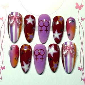 Pode incluir: Um conjunto de dez unhas artificiais em vários designs. As unhas apresentam uma mistura de cores, incluindo rosa, roxo e vermelho. Algumas unhas têm desenhos de estrelas e símbolos de gênero, outras padrões listrados e um laço.