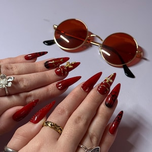Pode incluir: Unhas vermelhas em estilo stiletto com detalhes dourados e óculos de sol redondos tingidos de vermelho com armação dourada. As unhas apresentam desenhos e enfeites elaborados.
