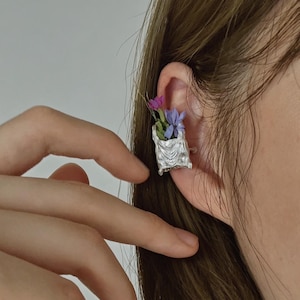 Op de afbeelding: Een zilverkleurige oormanchet met een getextureerde houder gevuld met paarse en roze bloemen. De manchet wordt op het oor gedragen en voegt een vleugje elegantie toe, geïnspireerd door de natuur. Het ontwerp is ingewikkeld en artistiek.
