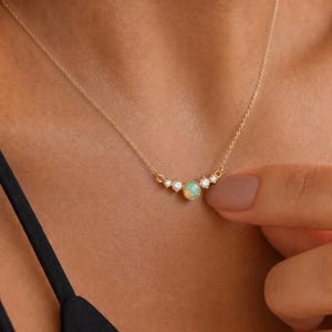 May include: A gold chain necklace featuring a central round opal gemstone surrounded by four smaller clear round stones. The pendant is set in a delicate gold frame with a beaded edge, suspended from a thin gold cable chain that rests on the collarbone.