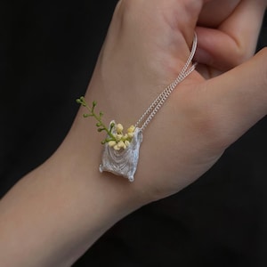 May include: A silver-toned pendant necklace featuring a small, textured pouch design. The pouch is filled with tiny yellow and green floral accents. The necklace is held in a hand against a dark background, showcasing the delicate details of the jewelry.
