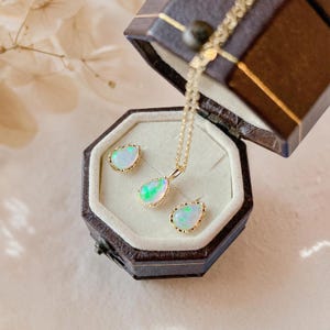 May include: A jewelry set featuring a gold necklace and two matching stud earrings, each adorned with a teardrop-shaped opal gemstone. The pieces are displayed inside a dark brown, hexagonal-shaped velvet-lined box with a hinged lid.