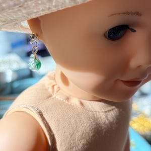 May include: A close-up of a doll wearing a straw hat and a silver earring. The earring has a small silver stud and a dangling teardrop-shaped charm with green and clear accents. The doll's skin is a light peach color.