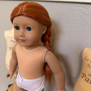 May include: A doll with red hair styled in a braid. The doll has blue eyes, a peach-colored top, and light pink underwear. The doll is standing in front of a neutral background.