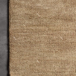 May include: Close-up of a rectangular, woven jute rug. The rug has a natural tan color and a textured surface. The bottom edge features a fringe of white fibers. The rug is likely used as a floor covering.