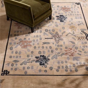 May include: A beige area rug with a floral pattern in shades of orange, gray, and black. The rug has a black border and a green armchair is partially visible in the upper left corner. The rug's design includes stylized flowers and leaf motifs.