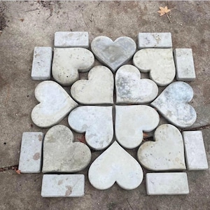 Puede incluir: Disposición de losas de hormigón en forma de corazón y bloques rectangulares. Las piedras son de color gris claro con un aspecto desgastado, dispuestas en forma de corazón y cuadrado. Ideal para senderos de jardín o paisajismo decorativo.