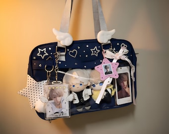 May include: A navy blue ita bag featuring a clear front pocket, two white plush wings on top, and grey shoulder straps. The bag is decorated with star and heart rhinestones, a large white polka-dot star charm, and two plush dolls inside.