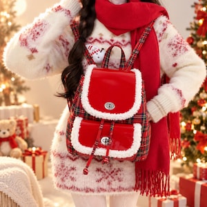 May include: A red and green plaid backpack featuring a glossy red flap and front pocket, both trimmed with white faux fur. The bag includes silver-tone metal twist-lock closures on the main compartment and the front pocket, with matching plaid straps.