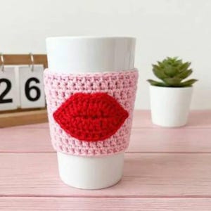 Peut inclure: Une tasse en céramique blanche avec une housse en crochet rose ornée d'un motif de lèvres rouges. Une petite plante succulente dans un pot blanc est à droite. La tasse est sur une surface en bois rose.