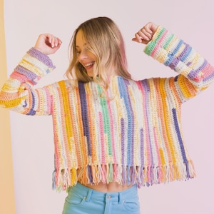 Pode incluir: Um top de crochê listrado colorido com decote em V e franjas. O top apresenta listras verticais em várias cores, incluindo amarelo, rosa, roxo e azul. As mangas são compridas e a bainha inferior tem franjas. O top é usado com jeans azul claro.