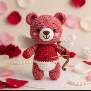 May include: A handmade crochet teddy bear in shades of red, with white wings and a bow and arrow. The bear has a white belly and a brown nose. The background includes rose petals and pearls, suggesting a Valentine's Day theme.