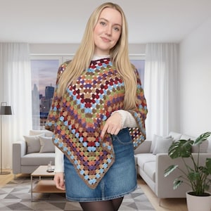 May include: A colorful, handmade crochet poncho with a square pattern in shades of brown, red, blue, green, and purple. The poncho is worn over a white long-sleeved shirt and a denim skirt. The garment has a bohemian style.