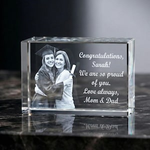 May include: A clear, rectangular crystal block with an engraved image of a graduate and a woman embracing. The text reads "Congratulations, Sarah! We are so proud of you. Love always, Mom & Dad."