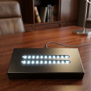 Op de afbeelding: Een zwarte, rechthoekige LED-lichtbasis met twee parallelle rijen helderwitte lampen. De basis staat op een houten bureau, met een leren stoel en boekenkasten op de achtergrond. Een lamp is ook zichtbaar.