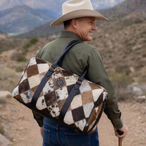 May include: A duffel bag featuring a diamond-patterned cowhide exterior in shades of brown, white, and black. The bag includes black leather handles, a top zipper closure, and a small rectangular leather patch with a logo on the front side.