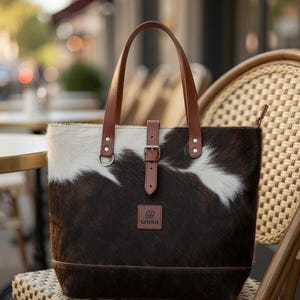 May include: A tote bag featuring a brown and white cowhide pattern with smooth brown leather handles and a central vertical strap with a metal buckle. A square brown leather patch with the embossed text LINDEN is centered on the front panel.
