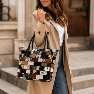 May include: A rectangular tote bag featuring a patchwork design of cowhide panels in shades of brown, black, white, and grey. The bag has two black leather handles attached to the top, a structured silhouette, and a textured, multi-colored finish.