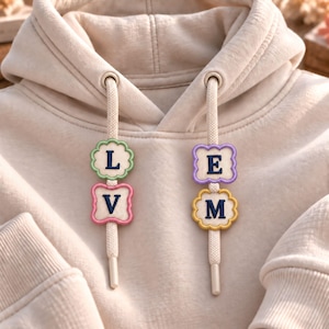 Puede incluir: Una sudadera con capucha de color beige que presenta dos cordones con cuatro parches decorativos de bordes festoneados. Los parches están bordados con las letras L, V, E y M en azul oscuro, enmarcadas por bordes verde claro, rosa, morado y amarillo.