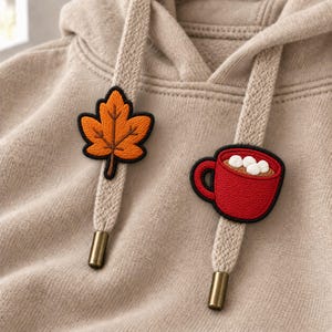 May include: A beige hooded sweatshirt featuring two decorative drawstring aglets. One drawstring has an embroidered orange maple leaf patch, while the other features an embroidered red mug filled with a brown beverage and white marshmallows.