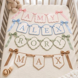 May include: Four decorative fabric banners featuring embroidered names AMY, ALEX, RORY, and MAX. Each banner consists of individual scalloped or shield-shaped panels connected by ribbons, with matching satin bows at the ends in pink, blue, sage, and tan.