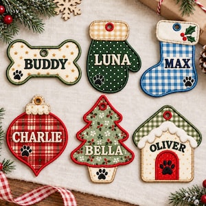 May include: Six fabric Christmas ornaments with embroidered names and paw prints. Shapes include a bone labeled BUDDY, a green mitten labeled LUNA, a blue plaid stocking labeled MAX, a red plaid bauble labeled CHARLIE, a tree labeled BELLA, and a house labeled OLIVER.
