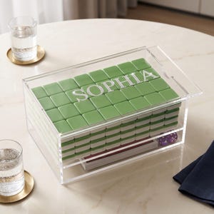 May include: A clear acrylic storage box containing a stack of green mahjong tiles. The top tile layer features the name SOPHIA in white, sparkling lettering. The box has a sliding lid and a lower compartment holding a deck of cards and purple dice.