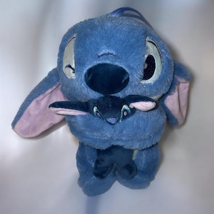 May include: A plush blue Stitch doll holding a smaller Stitch doll. The larger doll has large, expressive eyes, a dark blue nose, and large ears with pink insides. The smaller doll is dark blue with embroidered details.