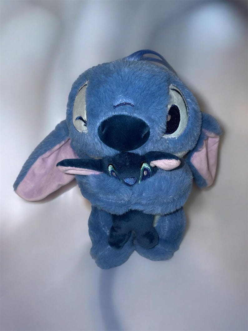 May include: A plush blue Stitch doll holding a smaller Stitch doll. The larger doll has large, expressive eyes, a dark blue nose, and large ears with pink insides. The smaller doll is dark blue with embroidered details.