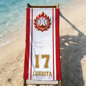 May include: A white beach towel with red stripes and a baseball-themed design. The design includes a baseball, stars, and flames. The number "17" and the name "LORISTA" are printed in gold.