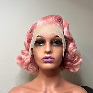 May include: A pink wig styled with vintage-inspired curls. The wig features a lace front and is displayed on a mannequin head with full makeup, including long eyelashes and purple lipstick. The wig's color is a soft, pastel pink.