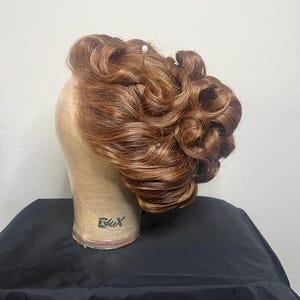 May include: A brown wig styled with voluminous, glossy curls, secured with a white pin. The wig is displayed on a mannequin head with a neutral skin tone, set against a plain background. The wig's color has highlights.