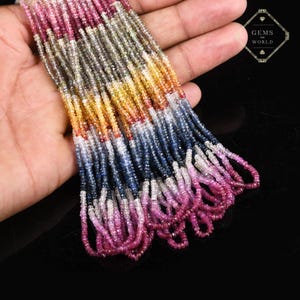 Könnte beinhalten: Eine Kette aus farbigen Edelsteinperlen in Rosa-, Blau-, Gelb- und Grautönen. Die Perlen sind klein und facettiert, aufgereiht. Das Bild enthält den Text "GEMS WORLD".