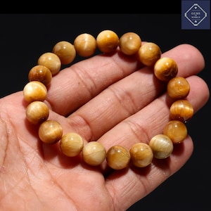 Op de afbeelding: Een gouden tijgeroog kralenarmband. De armband is gemaakt van ronde, gepolijste kralen met een chatoyante werking, waardoor een glinsterend, lichtreflecterend uiterlijk ontstaat. De kralen worden bij elkaar gehouden. De armband wordt in een hand gehouden.