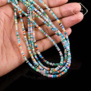May include: Strands of colorful gemstone beads in shades of blue, green, orange, and pink. The beads are small and round, strung together to create necklaces. The gemstones are held in a hand against a black background. The image has a Gems World logo.