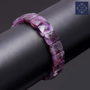 May include: A faceted amethyst bracelet displayed on a black stand. The bracelet features square-cut amethyst stones in varying shades of purple. The Gems World logo is visible in the upper right corner.
