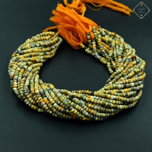 Pode incluir: Um fio de pequenas contas de pedras preciosas multicoloridas, principalmente amarelas, cinzentas e pretas. As contas são enfiadas juntas, formando um colar ou uma pulseira. Uma borla laranja está presa no topo. O fundo é cinza escuro.
