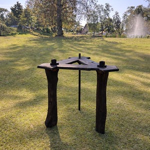 May include: A dark wooden table with a triangular top, supported by two tree trunk legs and a central metal pole. The table is set on a grassy lawn, with trees and a building in the background.