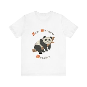 Puede incluir: Camiseta blanca con una ilustración de un oso panda. El panda está descansando en una rama, con las palabras "bear minimum monday" en una fuente divertida. El cuerpo del panda está hecho de texto.